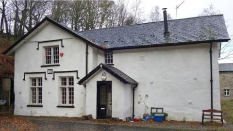 St Robert and St Alice Chapel at Dodding Green. The building is white with black window sills and a slate roof. There is a central doorway under a pitched roof. To its left is a wing that stands proud of the main building and has three windows, two on the ground floor and one centrally above. There is also a small window on the first floor above the roof over the main door. The part of the building to the right of the door is a single white painted wall with no windows.