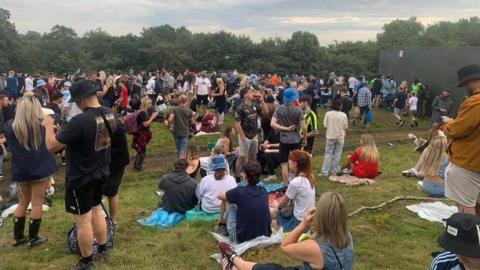 A grassy slope packed with a large crowd of people standing or sitting next to a tall fence. Trees can be seen in the background.