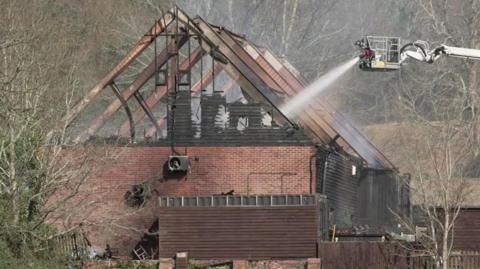Roof frame of barn building after fire - the roof is gone with just the frame left. Water is being sprayed into the barn from ladder platform by a fire crew.