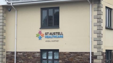 The exterior of a healthcare clinic building. The building has cream walls on the top half with a stone wall at the bottom. There are two windows and a logo next to text that says St Austell Healthcare. 