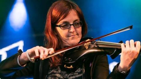 A woman with auburn hair, who is wearing glasses, playing the fiddle with blue concert lights in the background.