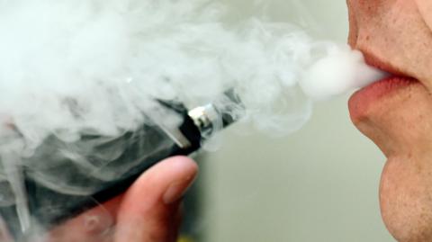 Close-up of a vape being held near lips. An anonymous person is vaping, blowing vapor out of their mouth.