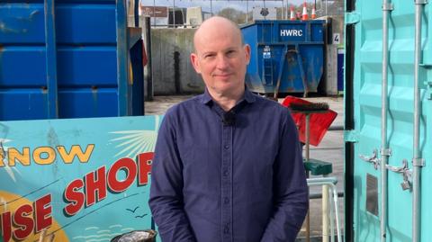 Loic has a bald head and wears a dark blue shirt, h stands outside the Re-use shop at Truro with a blue skip and cyan shipping container behind him. 