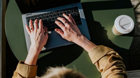 An aerial shot of a woman using a laptop computer. Her hands and the keyboards can be seen and she is wearing a brown shirt. 