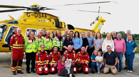 A group of 29 people and two babies. Half of the people are in every day clothes, standing and kneeling, while the others are wearing red flight suits- some with hi-vis vests. They are all in front of a yellow helicopter. 