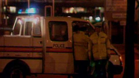 Night time, Police van with blue flashing light on top. A couple of police officers are visible getting out of the vehicle in high vis jackets and flat police hats.