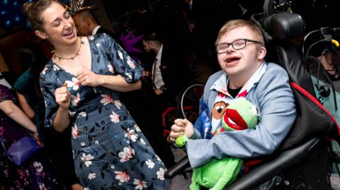 A woman in a floral dress is dancing with a boy in a wheelchair. He is wearing glasses and a light suit and is holding a Kermit the Frog toy. They are on a dance floor surrounded by other people.