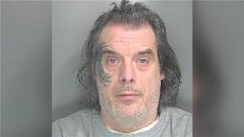 Lee White has shoulder length grey hair and a grey tattoo around his right eye. He's wearing a grey jumper, white-shirt and has blue eyes.