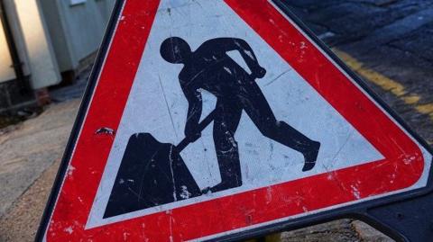 Generic image of a roadworks sign depicting a person with a shovel. There is a yellow line along the edge of a road in the background