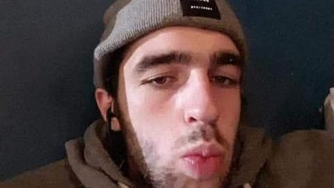 A selfie of Jamie Gilbey, a young man with dark hair and a beard wearing a beige beanie and brown hoodie while pouting. Behind him is a dark wall