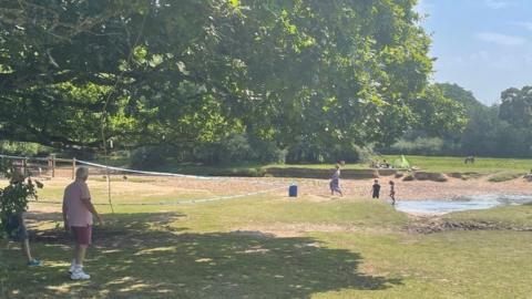 Cordon tape alongside a river with bathers and people sitting on the grass 