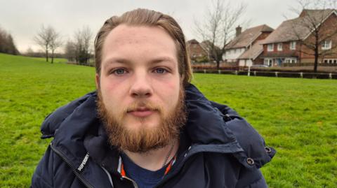 Sean Henry has a beard and slicked-back hair and is outside in a park with houses in the background. He is wearing a navy top and a navy outdoor jacket. There are trees with bare branches around the park.