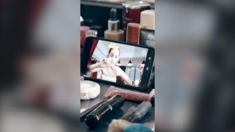 Young girl is seen sat in front of a phone screen that's recording her. She's pulling open a tube of makeup with her hands, and the phone is surrounded by various makeup products.