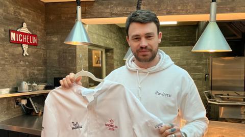 Liam Dillon is wearing a white hoody with the words, the boat written on the left side of his chest. He is holding a chef's uniform on a hanger and standing in a restaurant. On the wall there is the Michelin logo