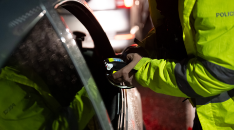 A police officer wearing hi-viz holds a breathalyser device through the driver's window of a car.
