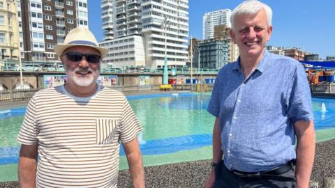 Two men are standing in front of a paddling pool, with blocks of flats in the background. The man on the left has a beard and sunglasses on, and wearing a striped white and brown t-shirt and a white hat. The man on the right has a blue short sleeved shirt and white hair