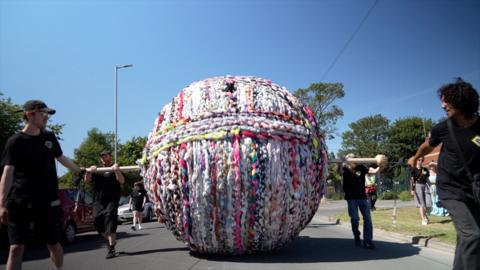 A giant ball of yarn is rolled along a road by people holding a piece of wood which runs through the ball.