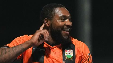 Britt Assombalonga in an amber and black Barnet FC jersey celebrates a goal during a match.