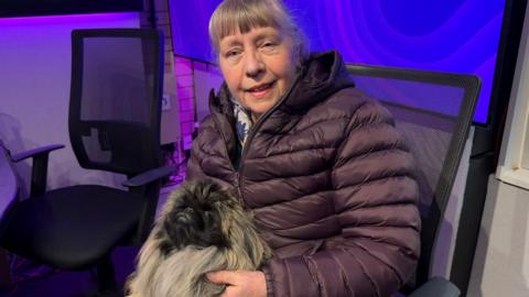 A woman with a grey full fringe, hair tied back and wearing a dark purple coat is sitting on a chair in a radio studio and holding a fluffy small dog in her lap. The dog has a black face and small nose with a fluffy head and body. It is pulling a side-eye expression on the camera as it sits in her lap.