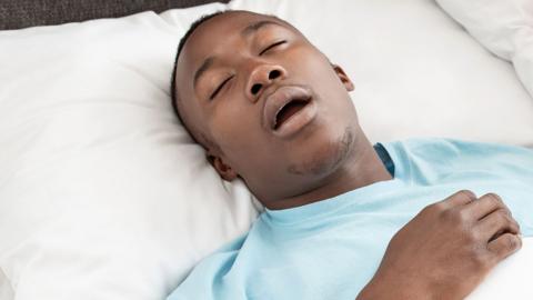 A young man is snoring while asleep on his back, his mouth open. He's wearing a light blue top and lying on top of a white pillow and white bed covers. 