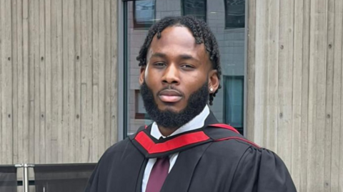 A man with short dreadlocks and a beard wearing a suit and a graduation gown.