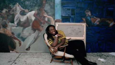 A man leans in a chair and grins while sitting in front of several large paintings.