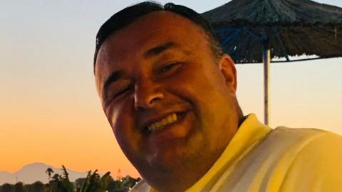 Photo of Paul Davinson, a man with a broad smile and yellow polo shirt, against the backdrop of a sunset and a parasol. 