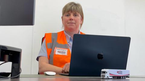 A woman wearing an orange high-vis vest sat behind a laptop with plain walls behind her.