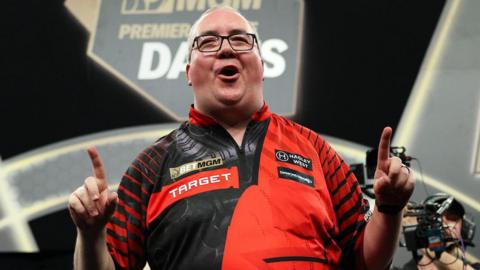 Stephen Bunting celebrates, pointing with both index fingers and pulling a celebratory facial pose as he wears a black and red darts shirt