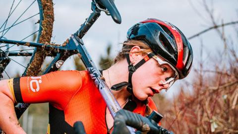 Lauren Fox is moving to the right, eyes ahead, carrying a mud-covered bike over her shoulder. She wears an orange cycling top and black and orange helmet. 