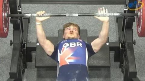 Boy with a blue and red gym shirt on with the letters "GBR" in bold white letters across and a Union Jack across his chest. He lies on a bench while lifting weights inside a gym