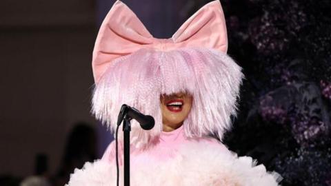 A woman wearing a large pink wig with a pink bow sings into a microphone
