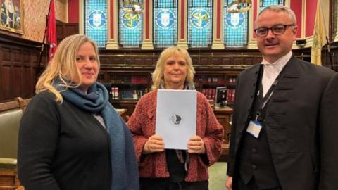 Michelle Heywood who has long light brown hair and is wearing a black cardigan and blue scarf, alongside Christa Viohl, who has shoulder length blonde hair  is wearing a salmon coloured cardigan and holding up the petition, and Juan Watterson who has close-cropped grey hair and is wearing glasses and is dressed in the Speaker's black jacket and waistcoat and white shirt, standing in House of Keys chamber.