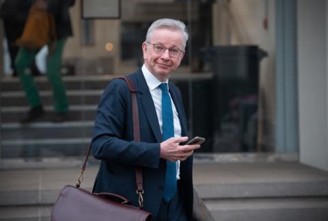 Michael Gove standing outside the Covid Inquiry on Monday, 10 March, after giving evidence. He is wearing a dark blue suit and staring straight at the camera while checking his mobile phone.