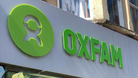 The sign above an Oxfam shop. The name and logo are cut from green plastic on a white background.