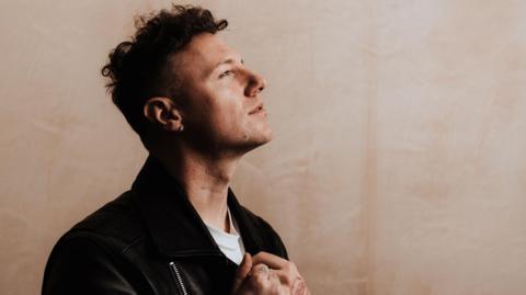 Callum Beattie has dark hair and is pictured in profile wearing a dark jacket over a white T-shirt. His head is titled upwards as he looks off to the distance.