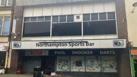 A view of the entrance to the Northampton Sports Bar. The windows of the former bar are covered in graffiti and there is some rubbish outside the door. Windows above the entrance have been blacked out. 