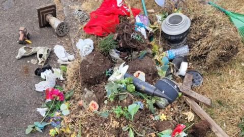 The contents of a bin, including broken ornaments, vases, graveside flower holders, grass, plants and soil, are strewn about by the side of a gravel path.