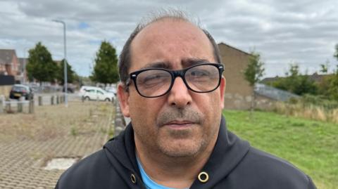 A man in a black hooded top, with a blue t-shirt underneath with black glasses looks straight at the camera. He is stood on a street with a house and cars in the background.