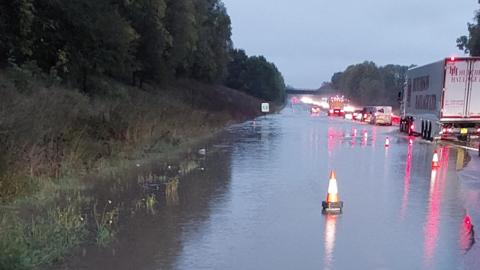 The A1 between Sedgefield and Newton Aycliffe. There is a lot of flood water and an orange cone is placed on the road. There are several vehicles, including lorries, vans and several cars.