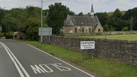 The A689 going through Eastgate.  It is a two-lane road with road markings reading slow. To the right, there is a sign reading Eastgate Please Drive Carefully.