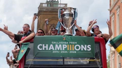 Rugby players celebrate with a huge silver trophy on an open-top red double-decker bus