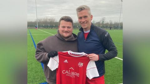 Jay Gurney and Steve Morison pose holding a red and white Hartwell Forest FC shirt. There is an all-weather pitch beside them.