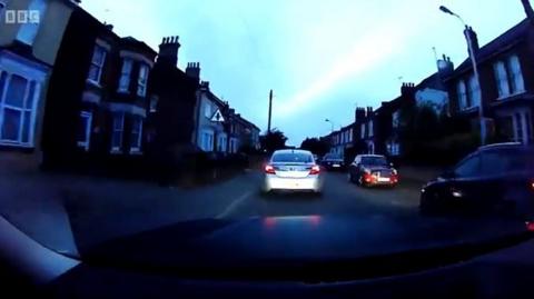 A still photo from a police pursuit video taken from the front window of the police car. A silver car can be seen driving through a housing estate at nighttime.