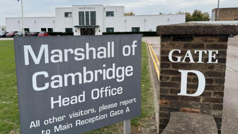 A large grey sign in the foreground says "Marshall of Cambridge Head Office". It is next to a brick pillar with "Gate D" on it. In the background is the company's white Art Deco reception building.