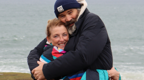 Julie and Greg hug on a beach while dressed for the cold weather