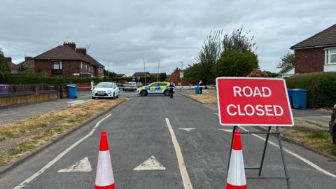 Image shows cones and a 'Road Closed' sign blocking a residential street in Huyton, Merseyside. 