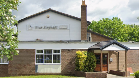 A brick building with a white-clad second floor an a covered porch leading to a brown wooden door