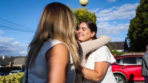 A woman with long blonde hair stands in front of two women hugging each other.