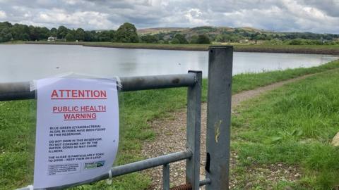 The reservoir surrounded by green grass and with a "public health warning" sign taped to a steel fence or gate